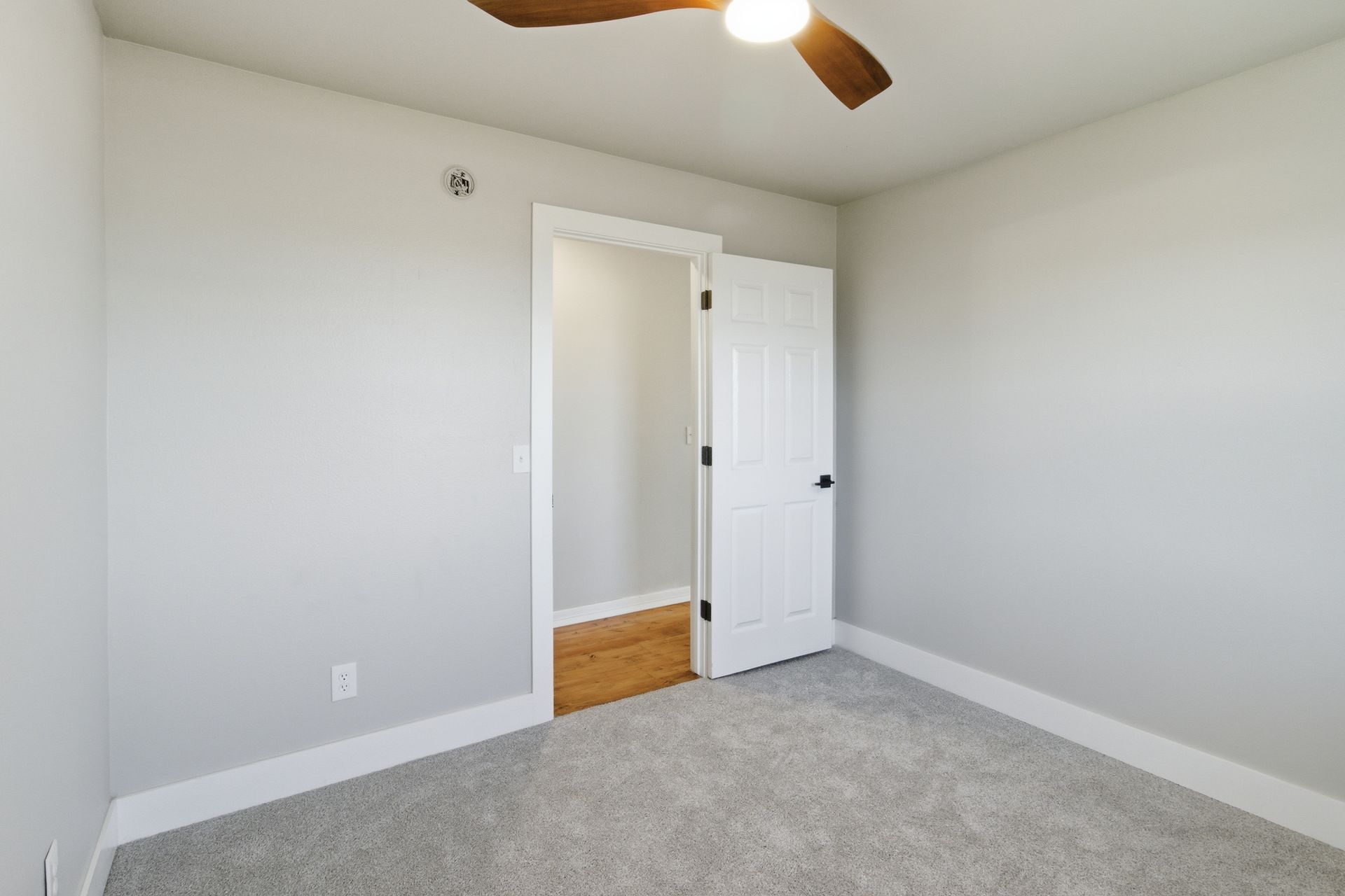 An empty room with light gray walls, gray carpet, and an open white door leading to a space with wood flooring.
