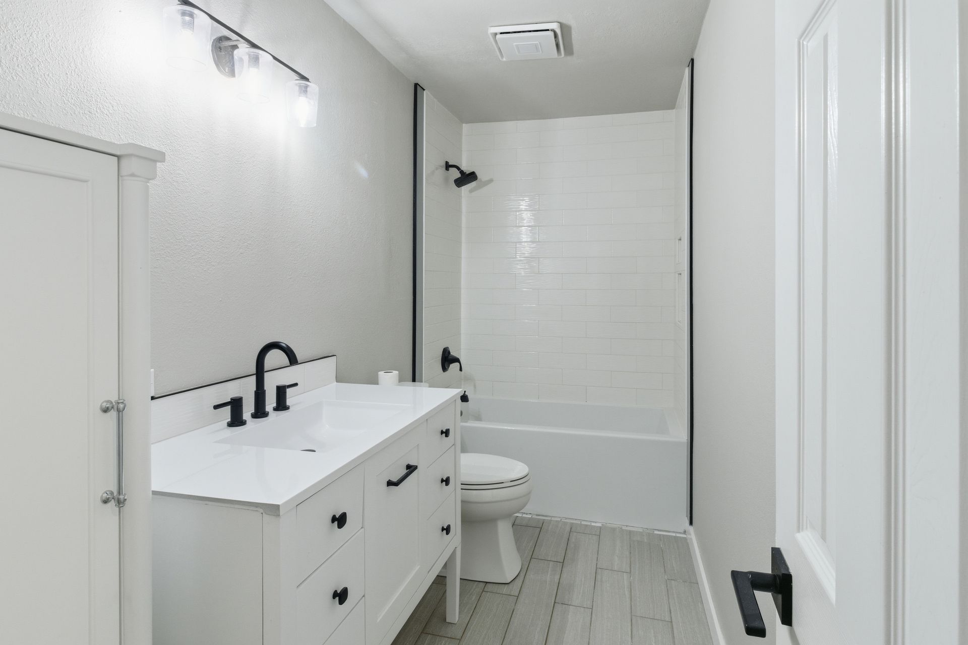 A bright, modern bathroom featuring a white vanity with black hardware, a toilet, and a bathtub with white tile walls.