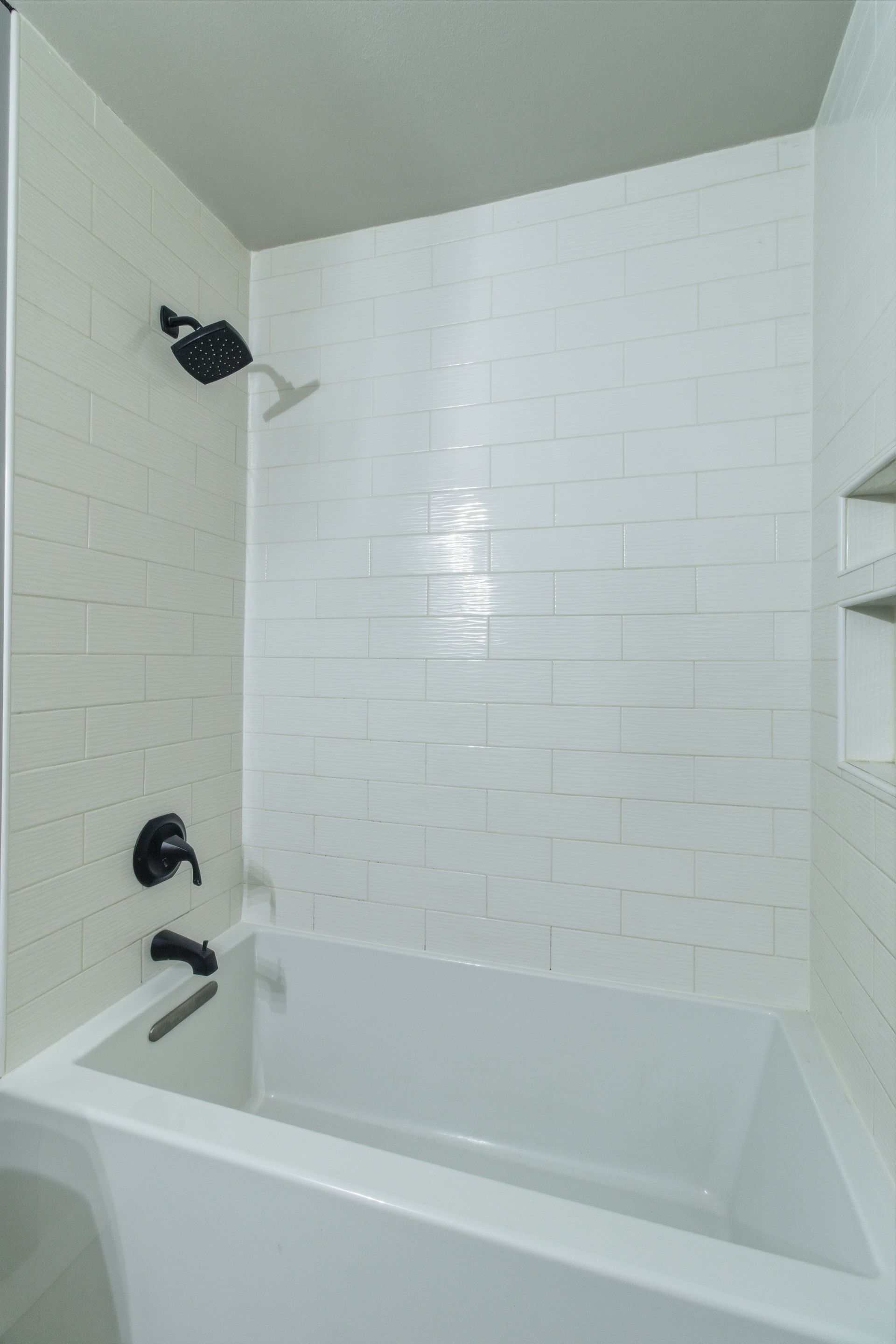 A white bathtub with a modern, white tiled wall, a shower head, and black fixtures in a contemporary bathroom.