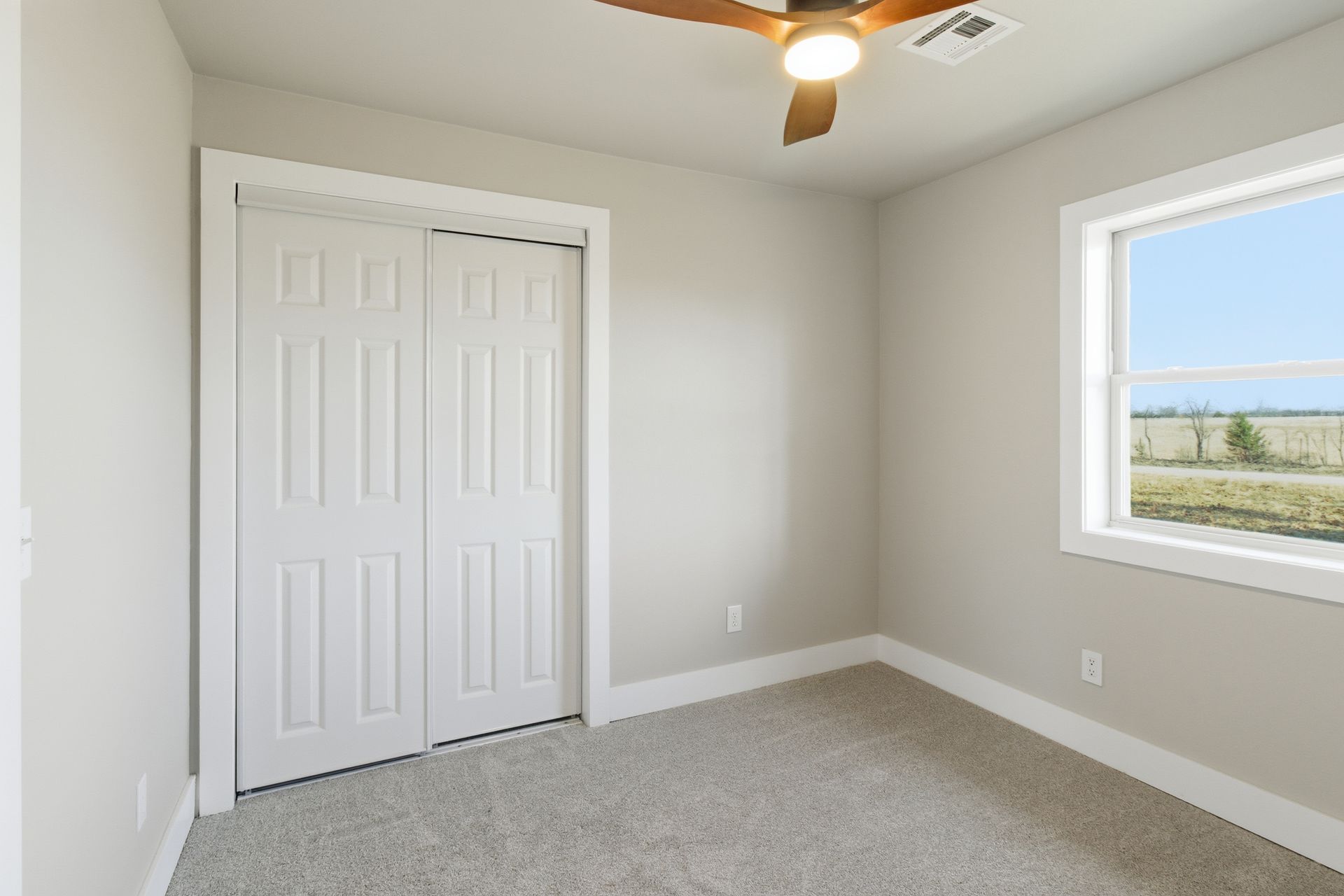 An empty bedroom with light gray walls, gray carpet, a white two-panel closet door, and a window with a rural view.
