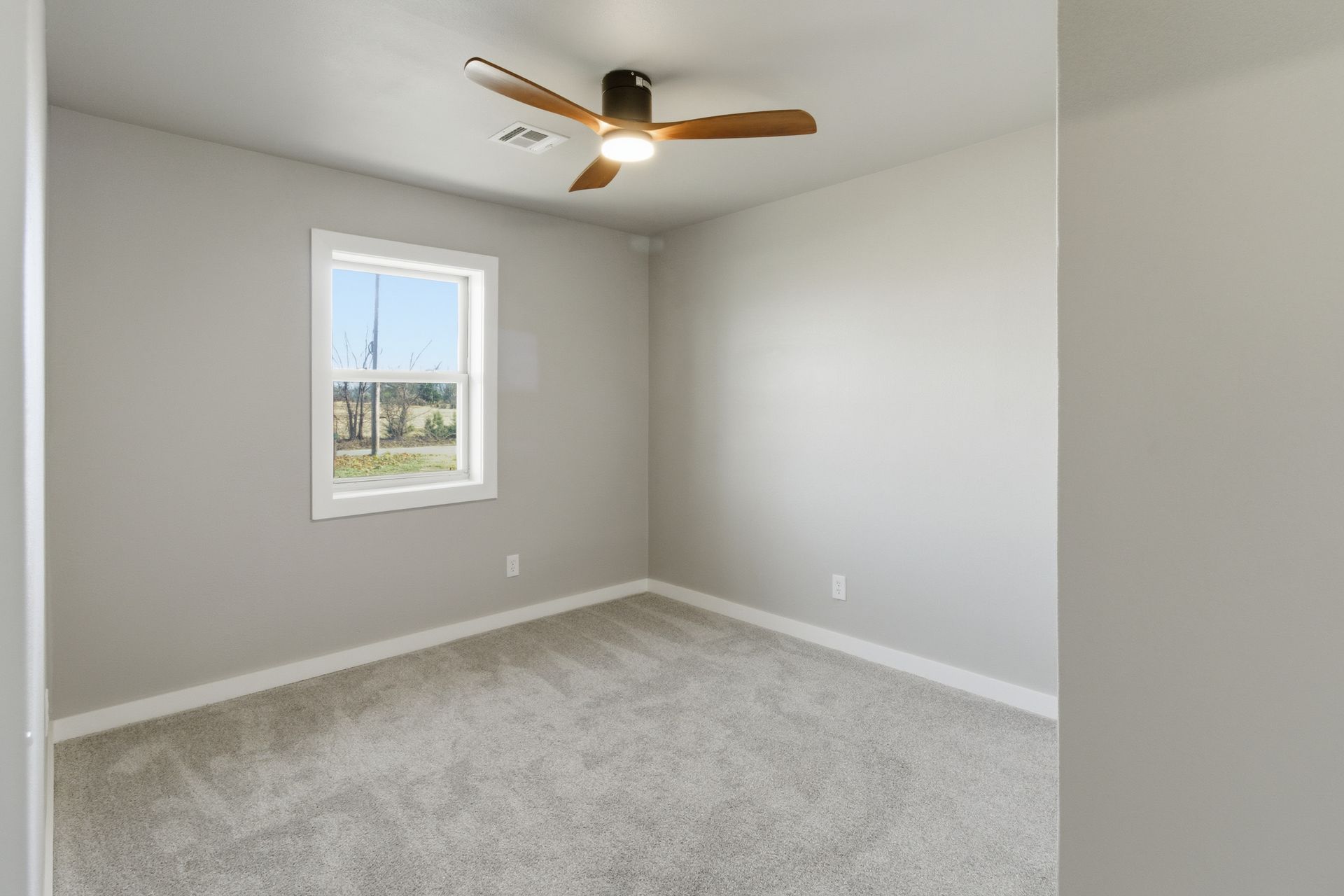 An empty room with light gray walls, gray carpet, a ceiling fan with wooden blades, and a single window.