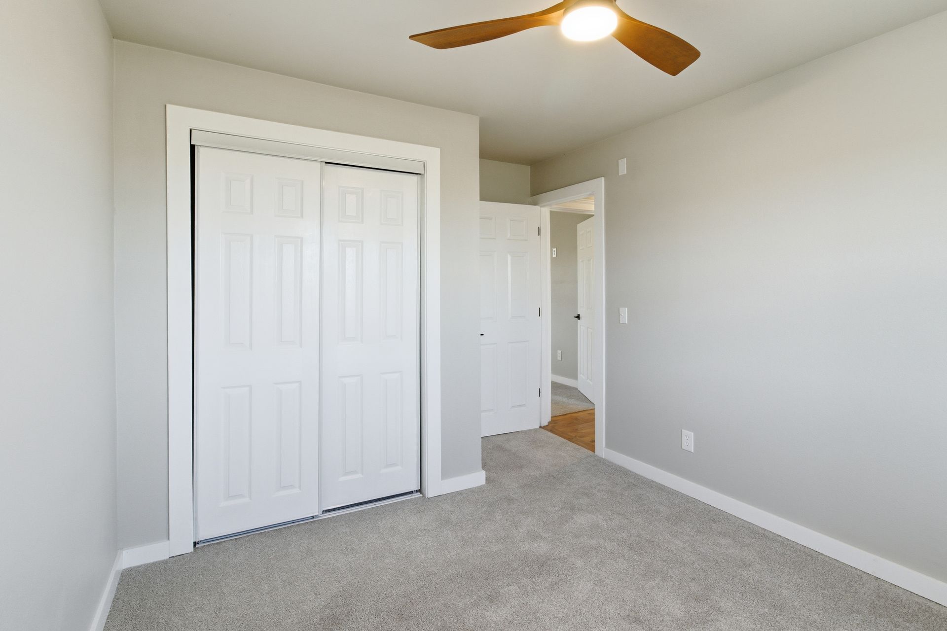 A simple, bright bedroom with light gray walls, gray carpeting, white closet doors, and a ceiling fan.