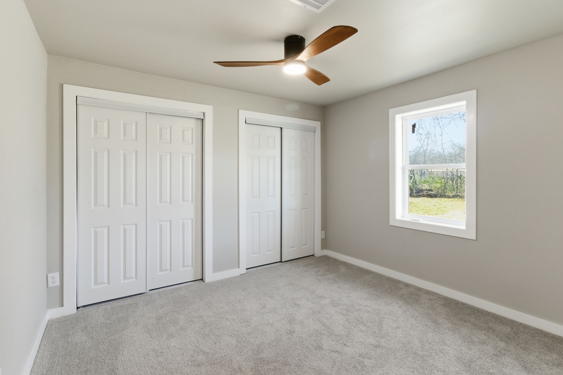 Empty bedroom with light gray walls, gray carpet, two white paneled closet doors, and a ceiling fan.