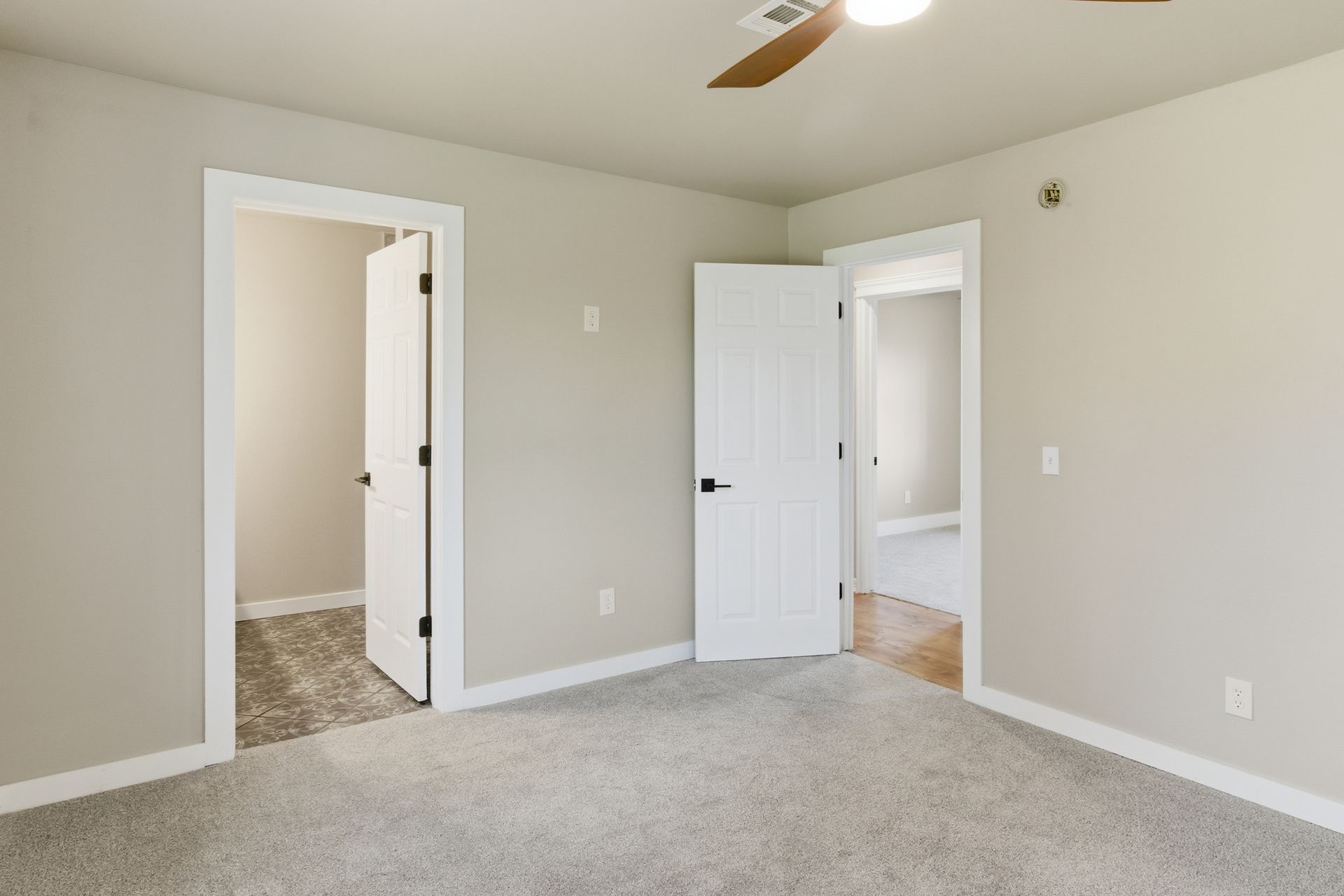 A bedroom interior with beige walls, grey carpeting, two open doorways with white doors, and a ceiling fan.