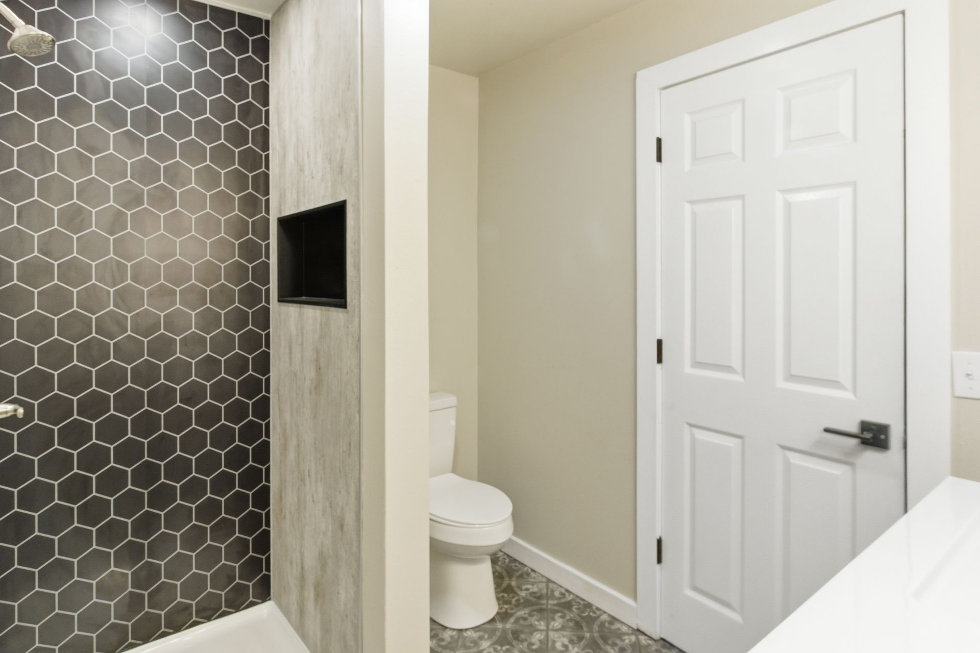 A modern bathroom interior featuring a grey hexagon-tiled shower, a white door, and a toilet in a separate alcove.