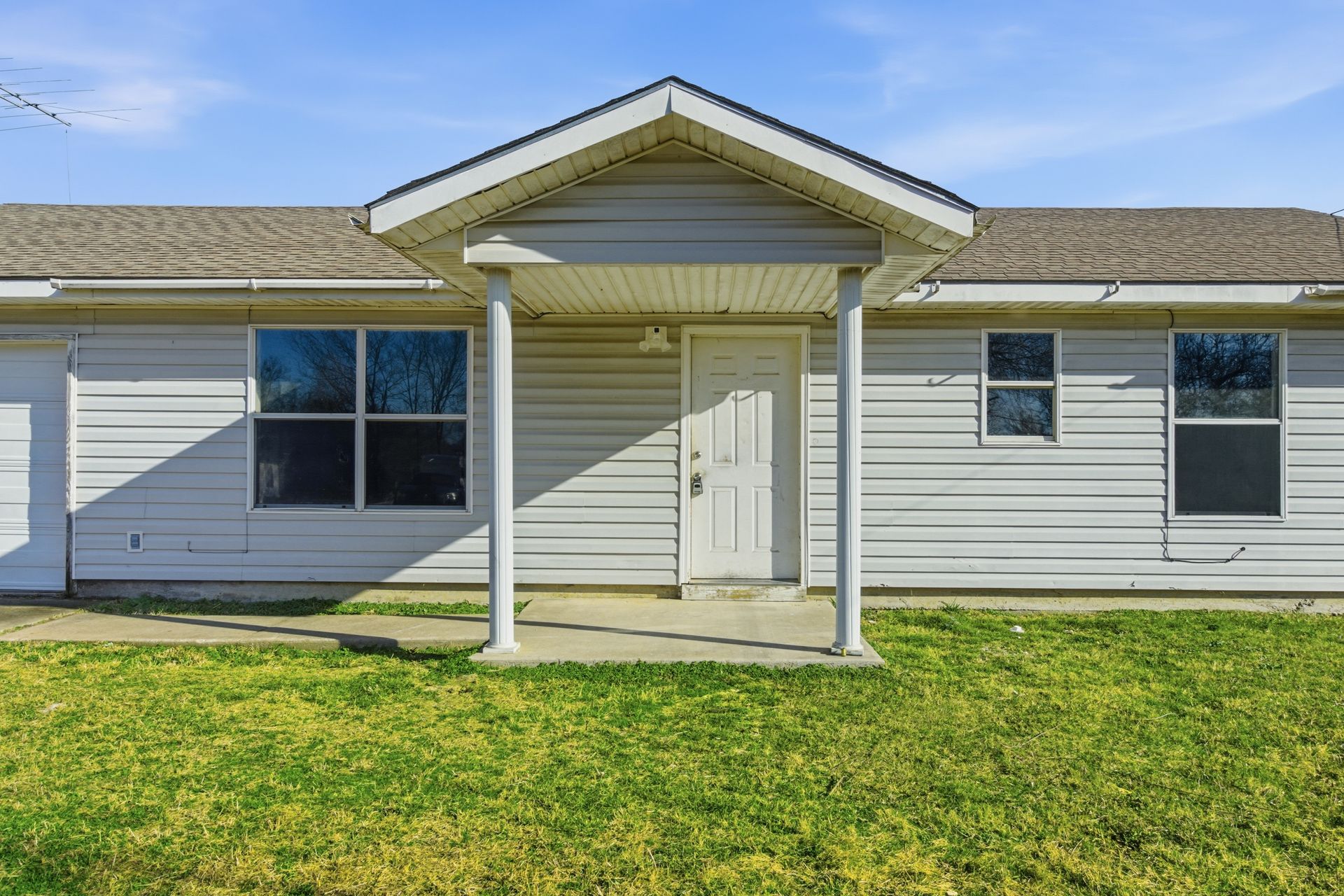 A single-story, light gray house with a covered front porch, white columns, and a green grass lawn under a blue sky.