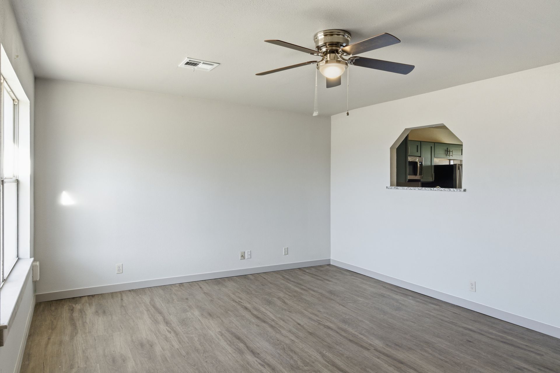 An unfurnished living room with white walls, wood-look flooring, a ceiling fan, and a pass-through window to the kitchen.