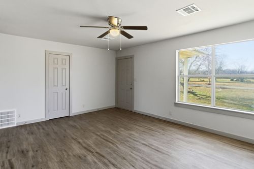 A minimalist, empty room with light gray walls, wood-look flooring, a ceiling fan, two doors, and a large window.