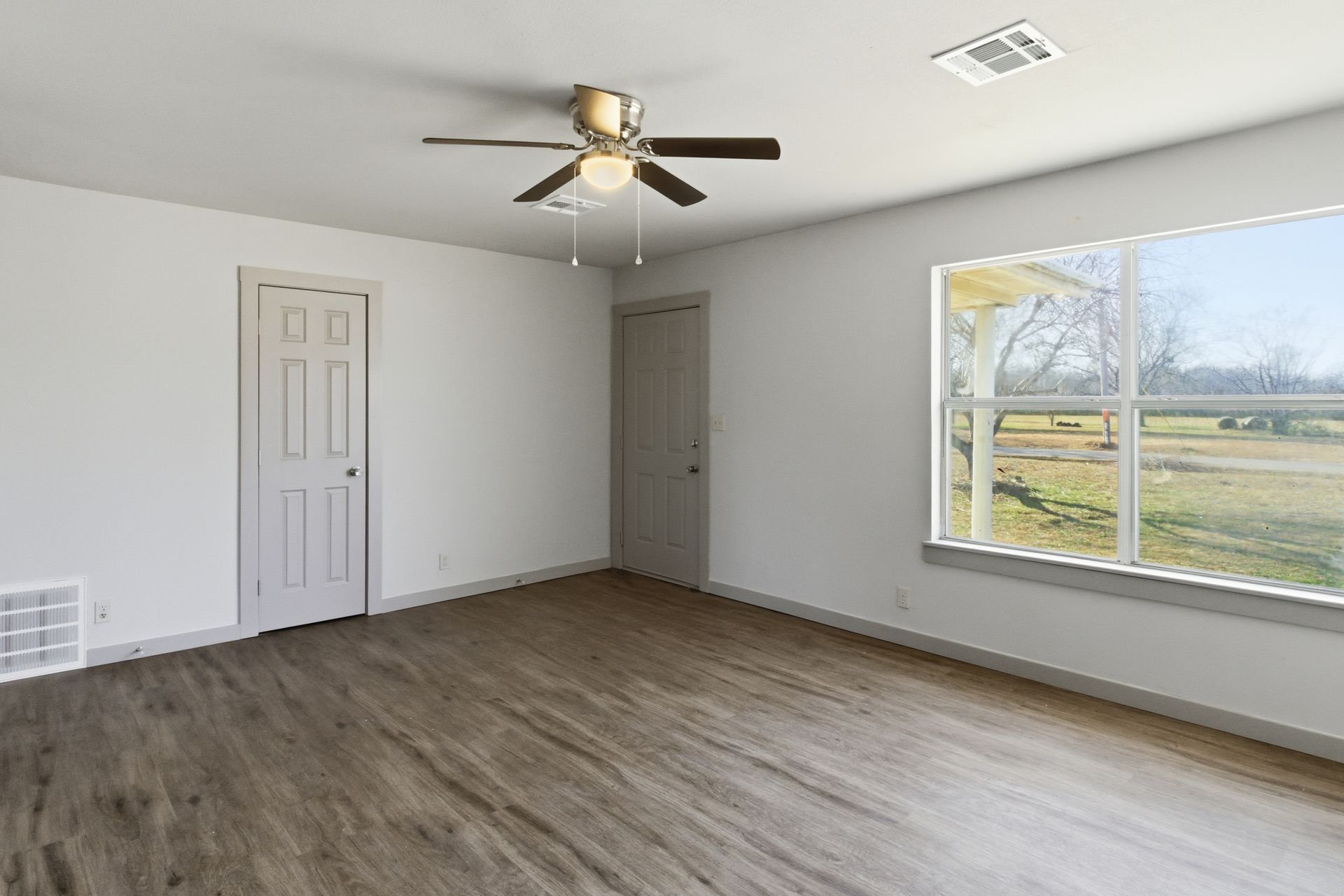 An empty bedroom with white walls, wood-look flooring, a ceiling fan, two closed doors, and a large window facing outside.