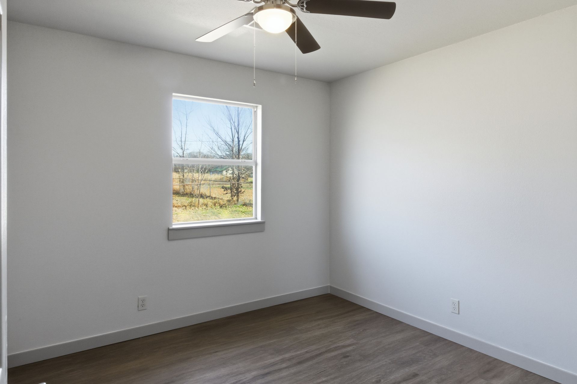 A bright, empty bedroom with white walls, wood-look flooring, a window showing a field, and a ceiling fan.