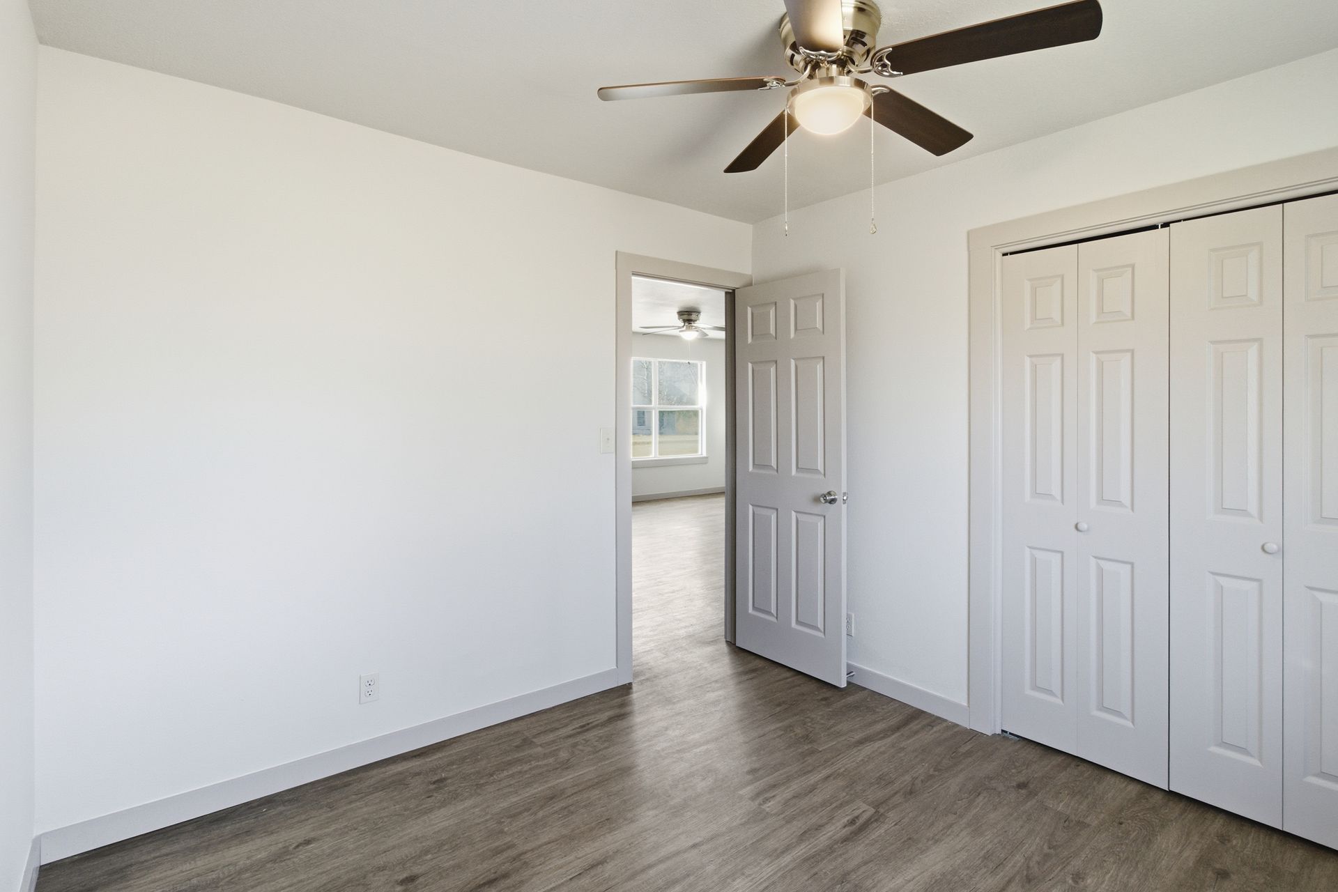 A modern, empty bedroom with white walls, wood-grain flooring, a ceiling fan, a doorway, and double closet doors.