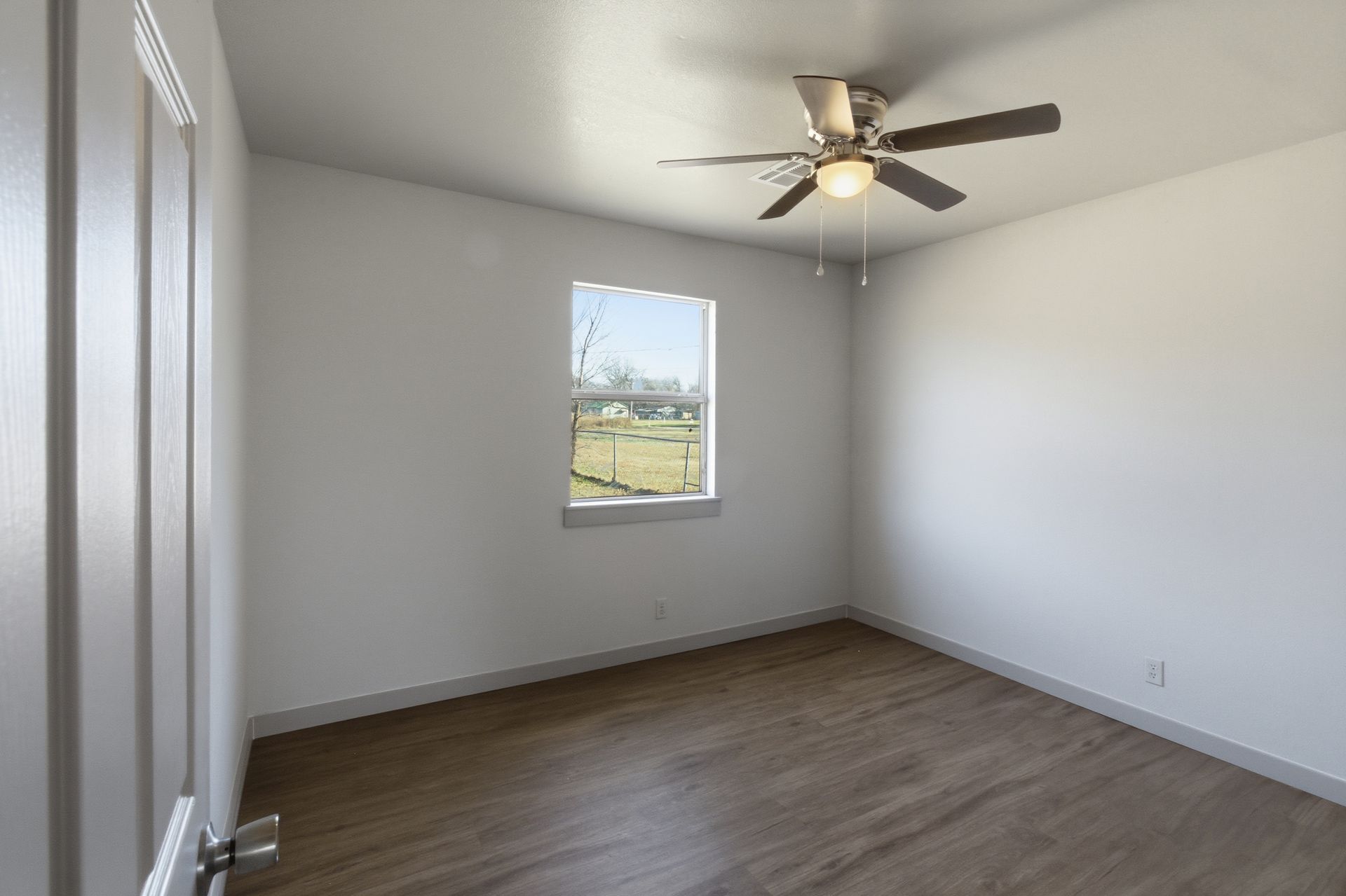 An empty bedroom with white walls, wood-look flooring, a ceiling fan, and a single window facing an outdoor landscape.