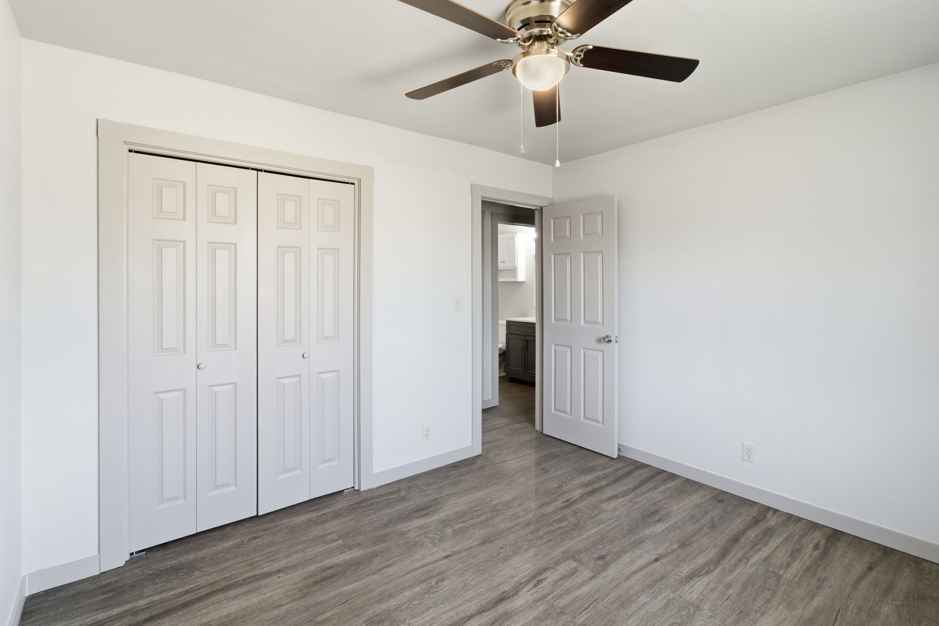 A bright, empty bedroom with white walls, gray wood-look flooring, a ceiling fan, a bifold closet, and a doorway.