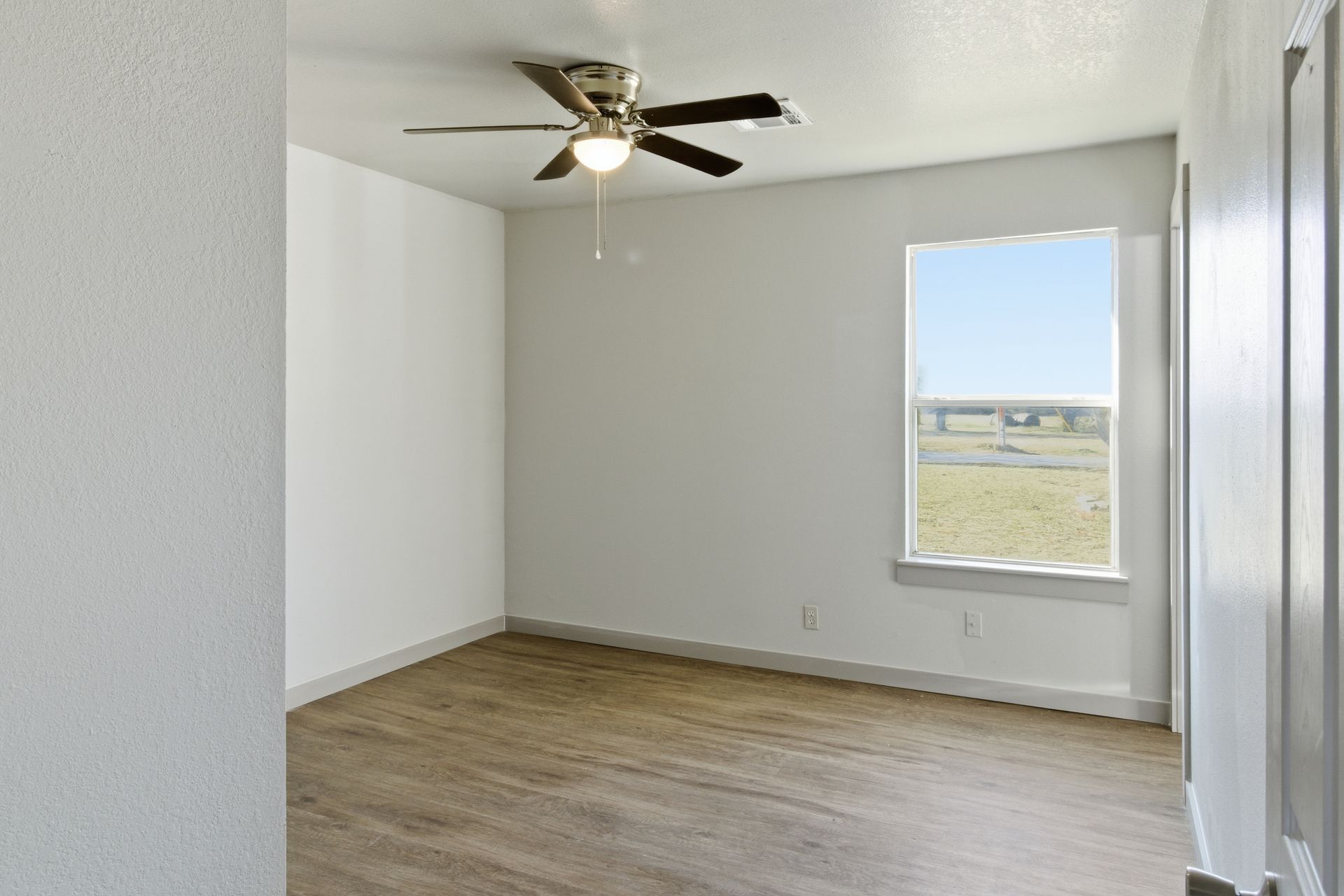 Empty room with white walls, wood-look flooring, a ceiling fan, and a single window looking out over a grassy landscape.
