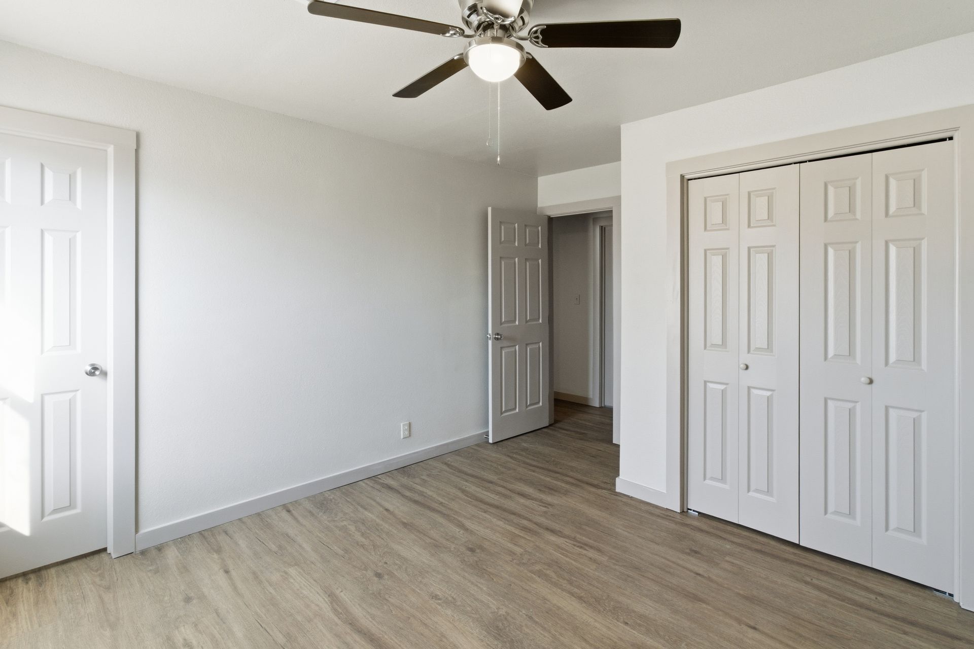 An empty, brightly lit bedroom with white walls, wood-look flooring, a ceiling fan, and white doors and closets.
