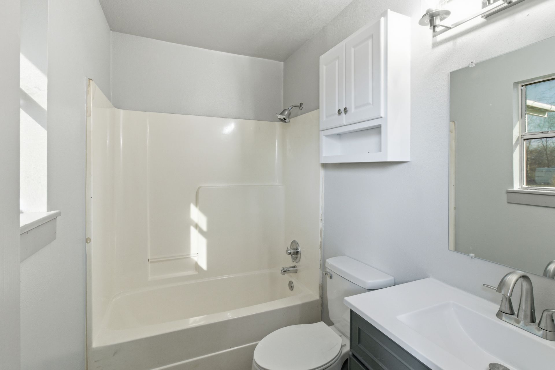 A modern bathroom featuring a white bathtub-shower combo, a white vanity with a gray cabinet, and a wall-mounted cabinet.