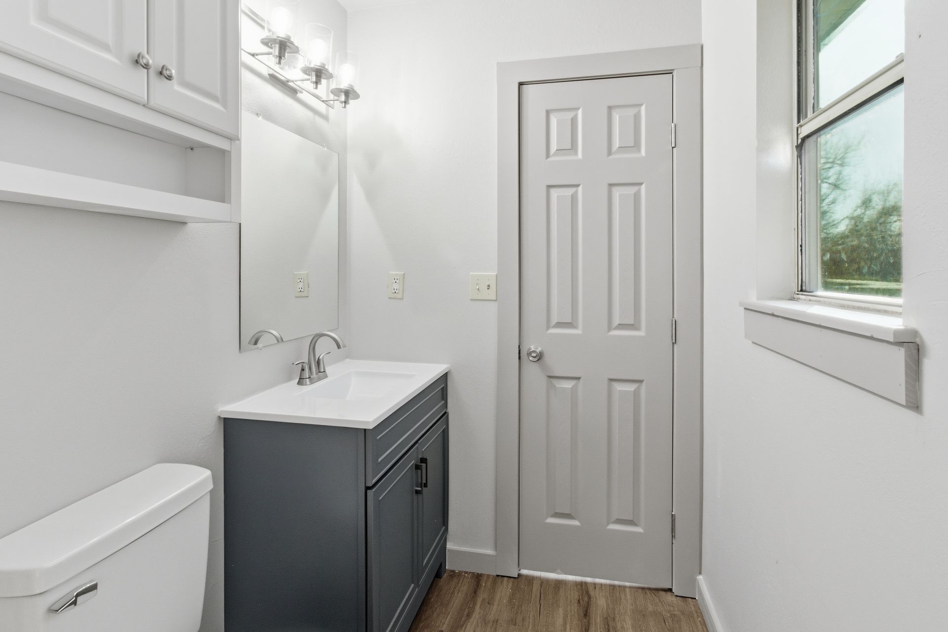 A bathroom interior with a grey vanity, white toilet, a mirror, and a white six-panel door near a window.