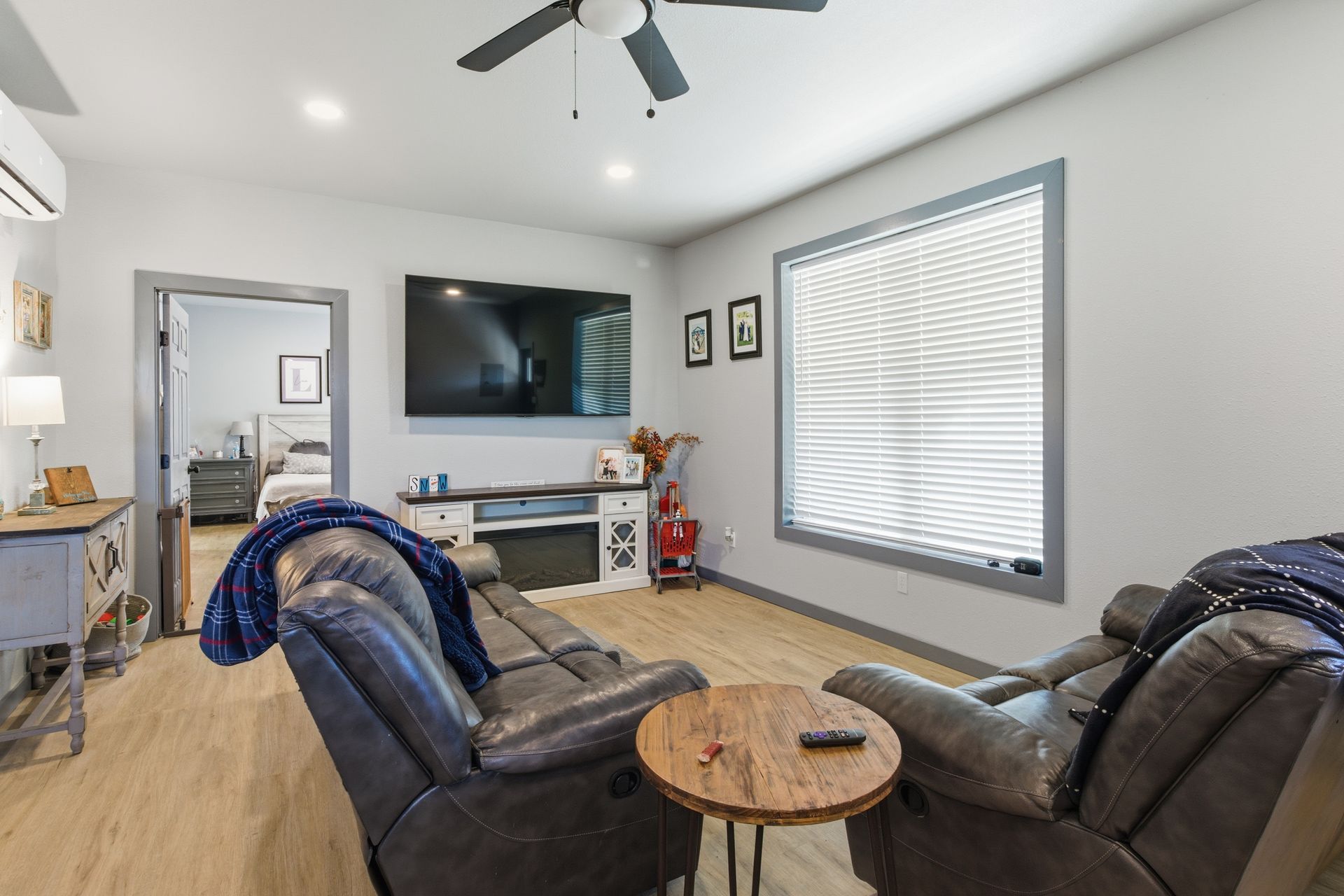 A living room with two gray recliners facing a TV, a wooden coffee table, light wood floors, and a view into a bedroom.