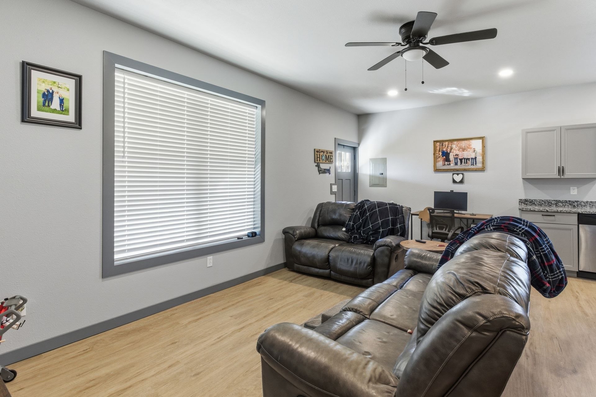 A living room featuring light wood floors, a large window with blinds, two dark leather sofas, and a workspace.