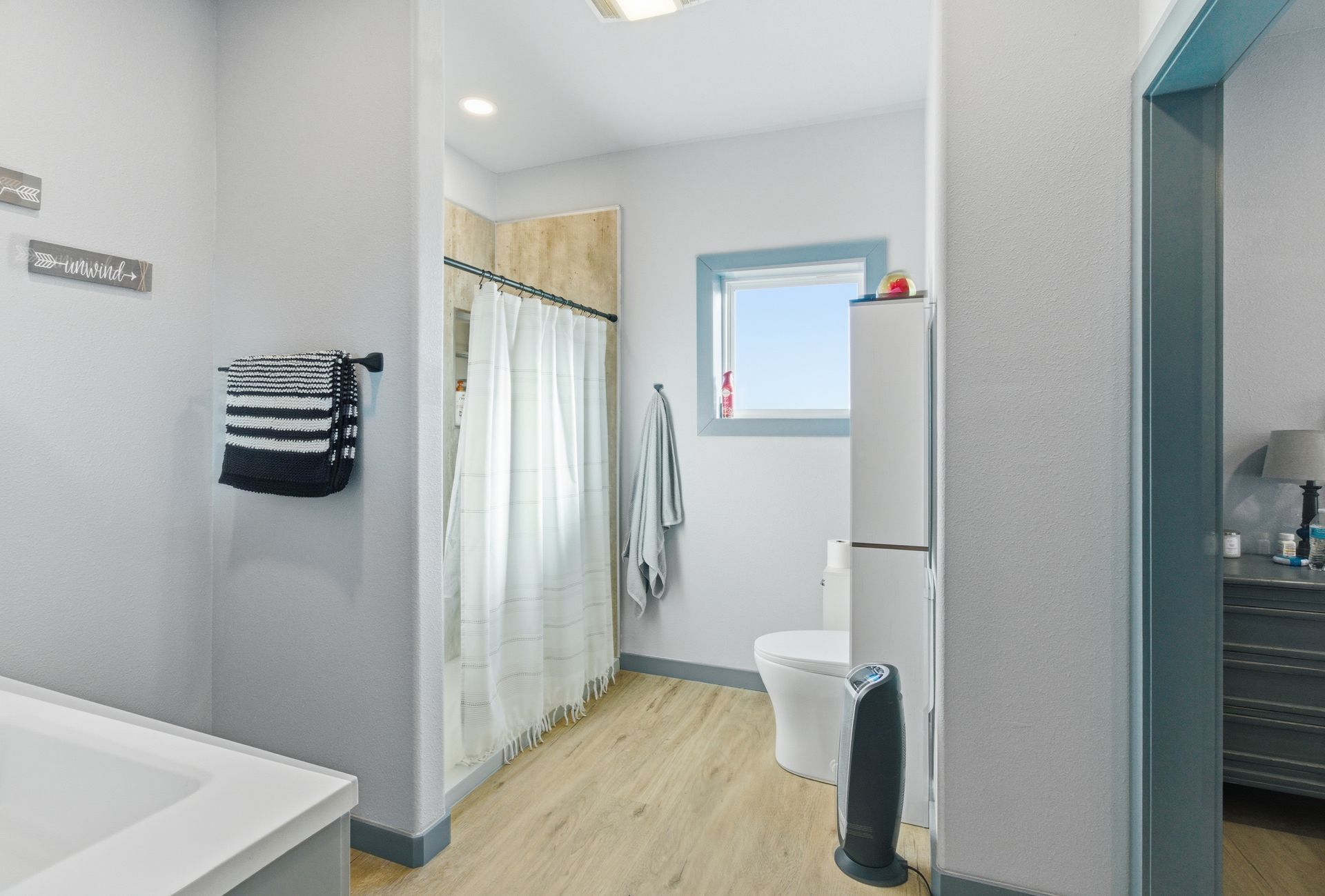 A bright bathroom with light wood floors, a white shower curtain, a toilet, and a window with a blue frame.