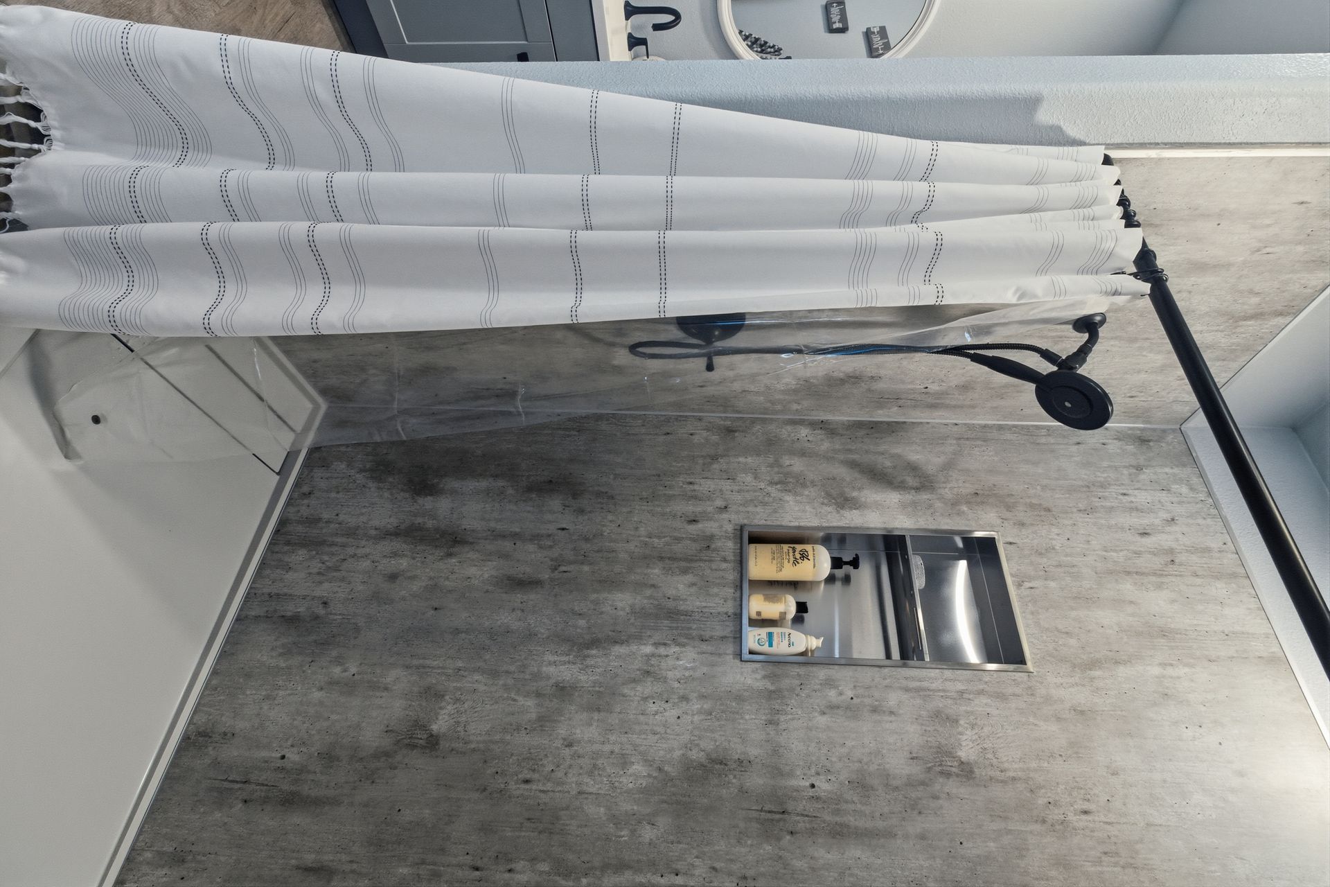 A modern shower with concrete-patterned tile, a recessed shelf containing toiletries, and a white, fringed shower curtain.