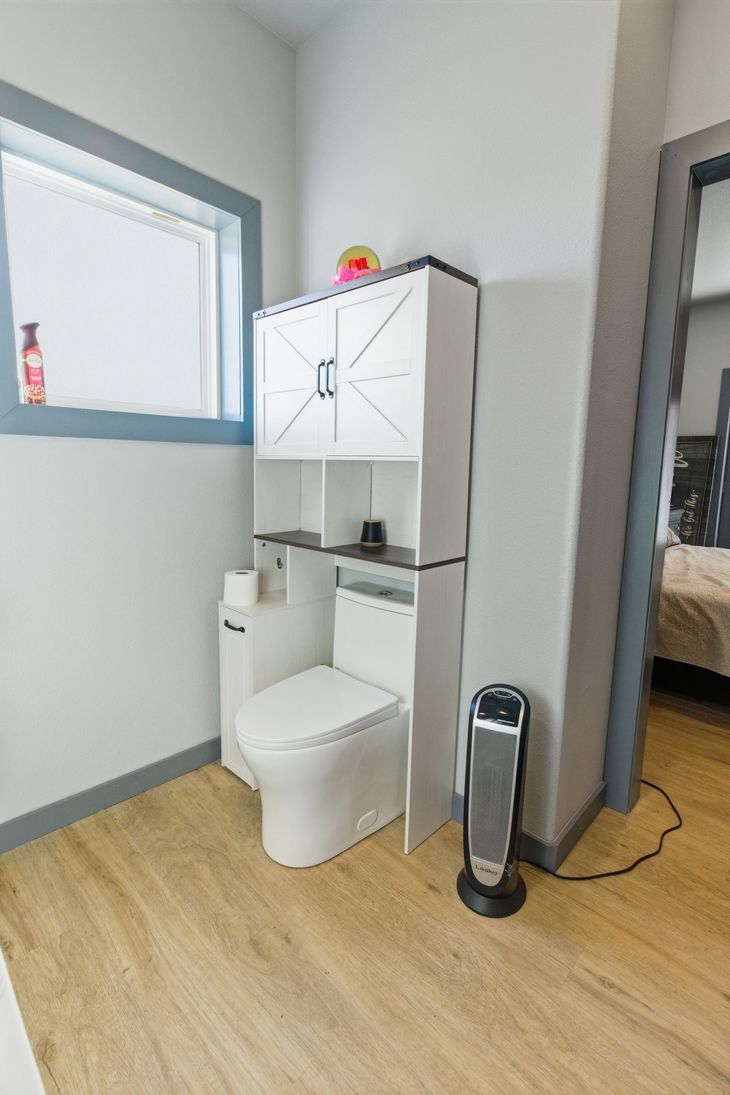 A white toilet with an over-the-toilet storage cabinet in a room with light wood floors, a gray-trimmed window, and a heater.