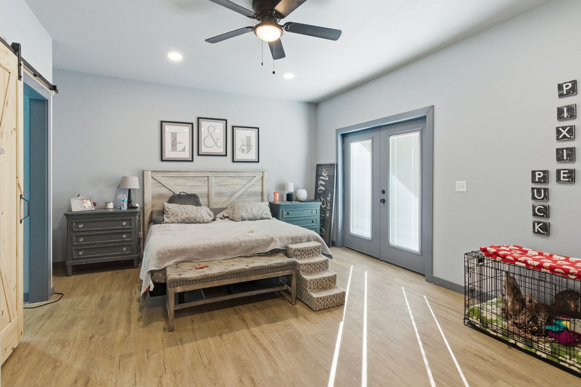 A bright, modern bedroom with wood-look floors, a wooden bed frame, a grey dresser, and a dog crate near double doors.