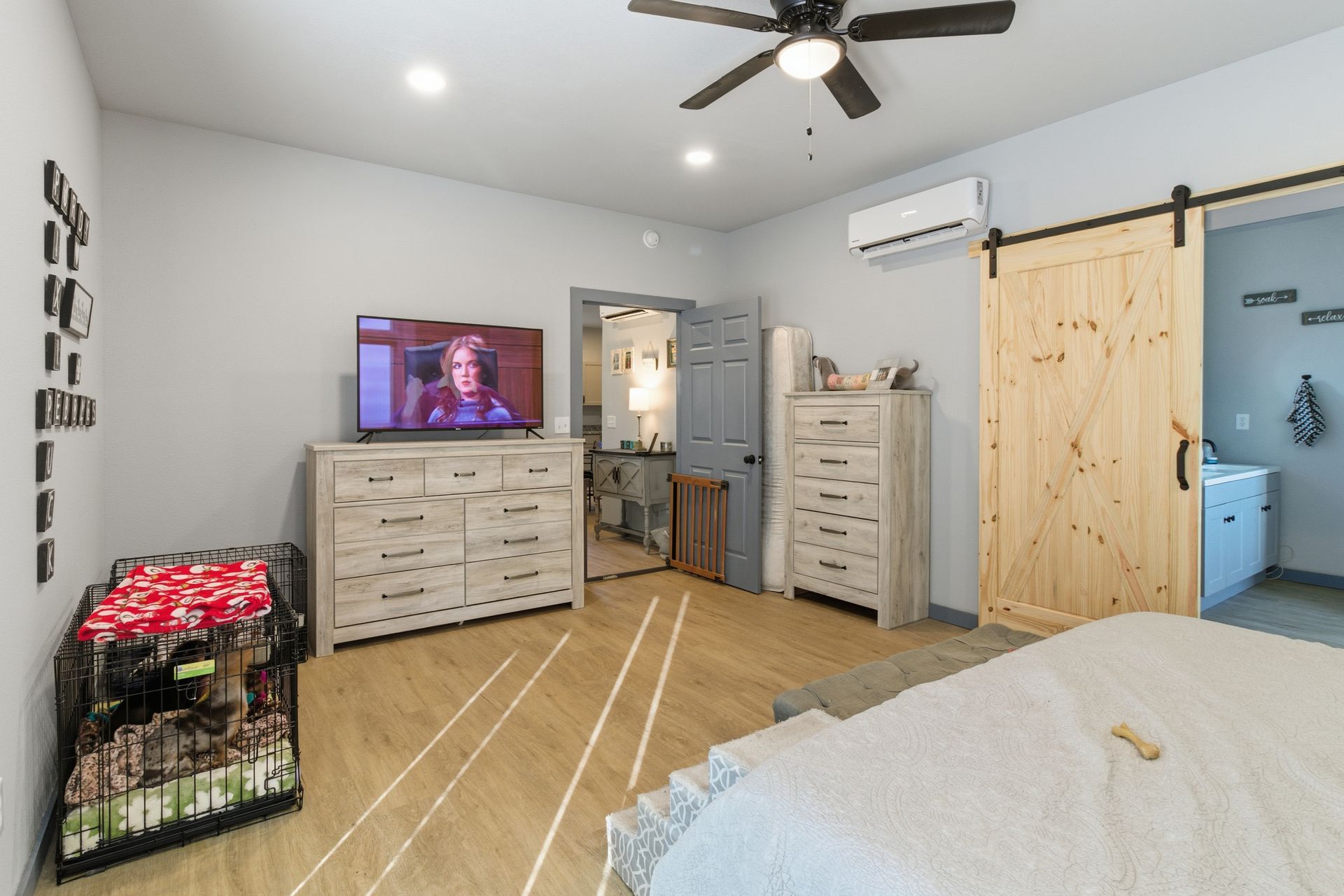 A light-colored bedroom featuring a television on a dresser, a dog crate, a ceiling fan, and a wood barn door to a bathroom.