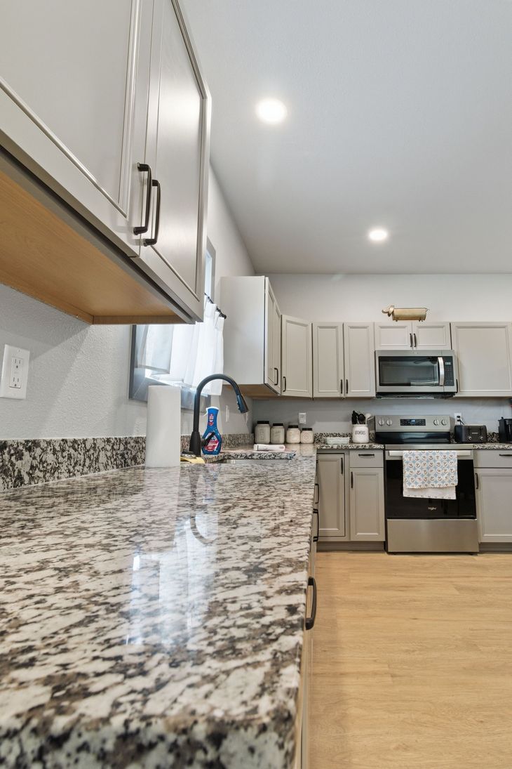 A modern kitchen with light gray cabinets, granite countertops, stainless steel appliances, and wood-look flooring.