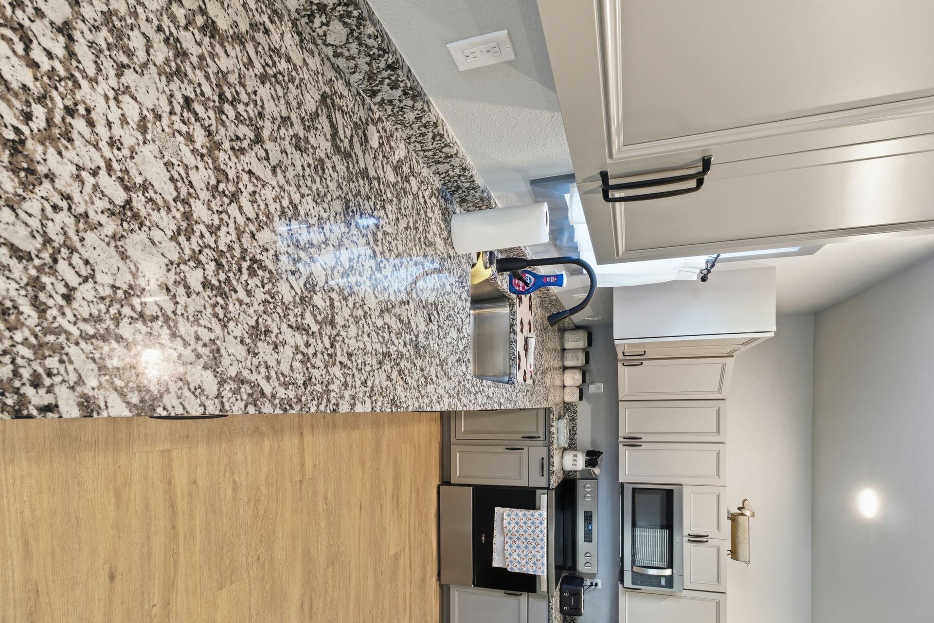 Modern kitchen with light grey cabinets, granite countertops, a stainless steel sink, and light wood-tone flooring.