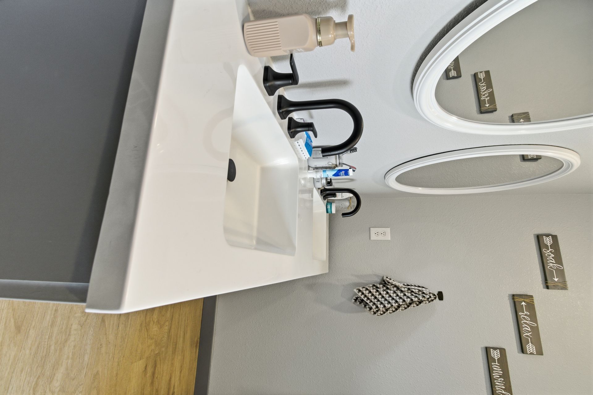 A modern bathroom vanity with a white countertop, black faucet, sink, and two oval mirrors on a light gray wall.