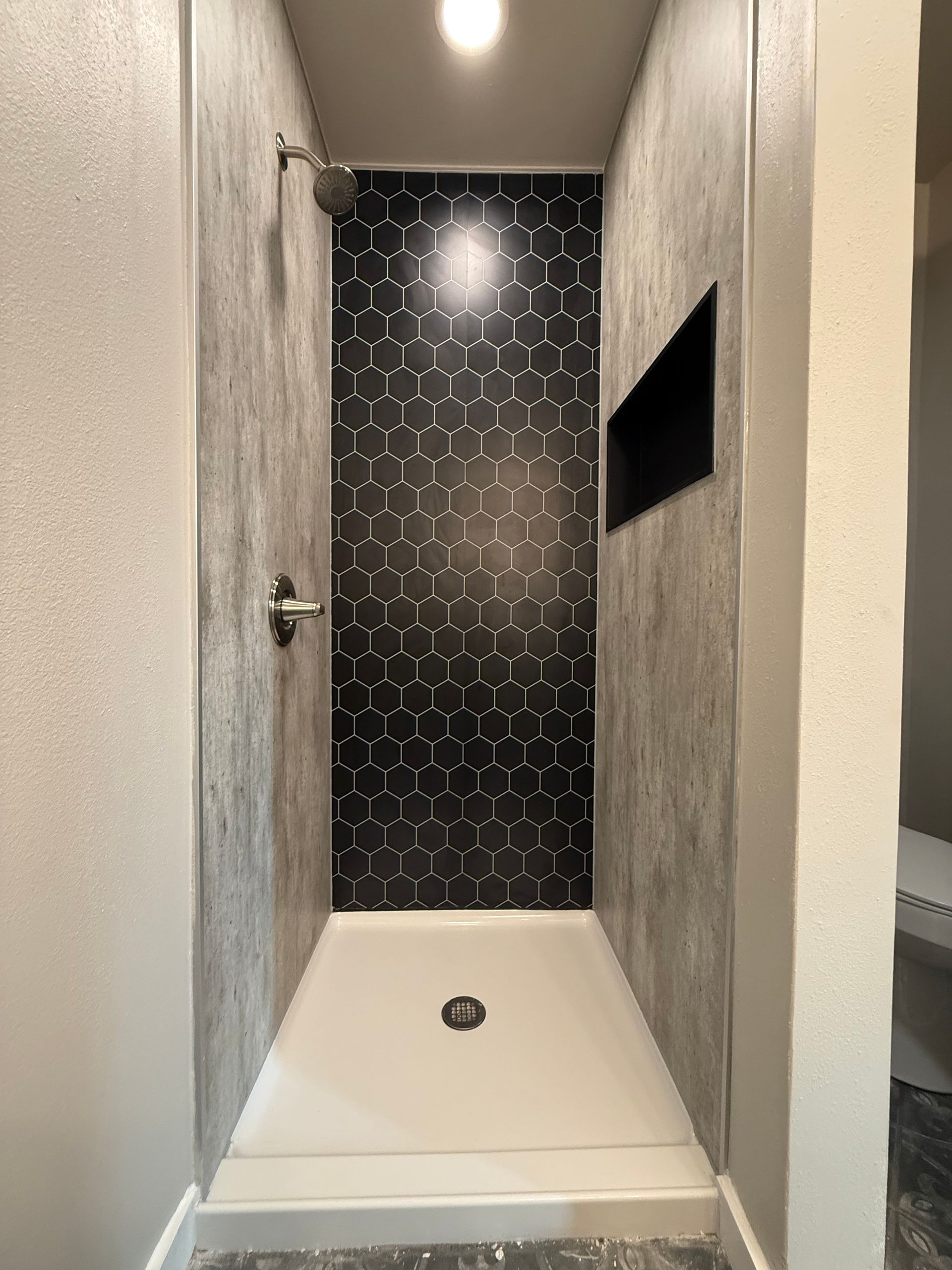 Narrow shower stall with white base, black hex tile back wall, and small shelf opening