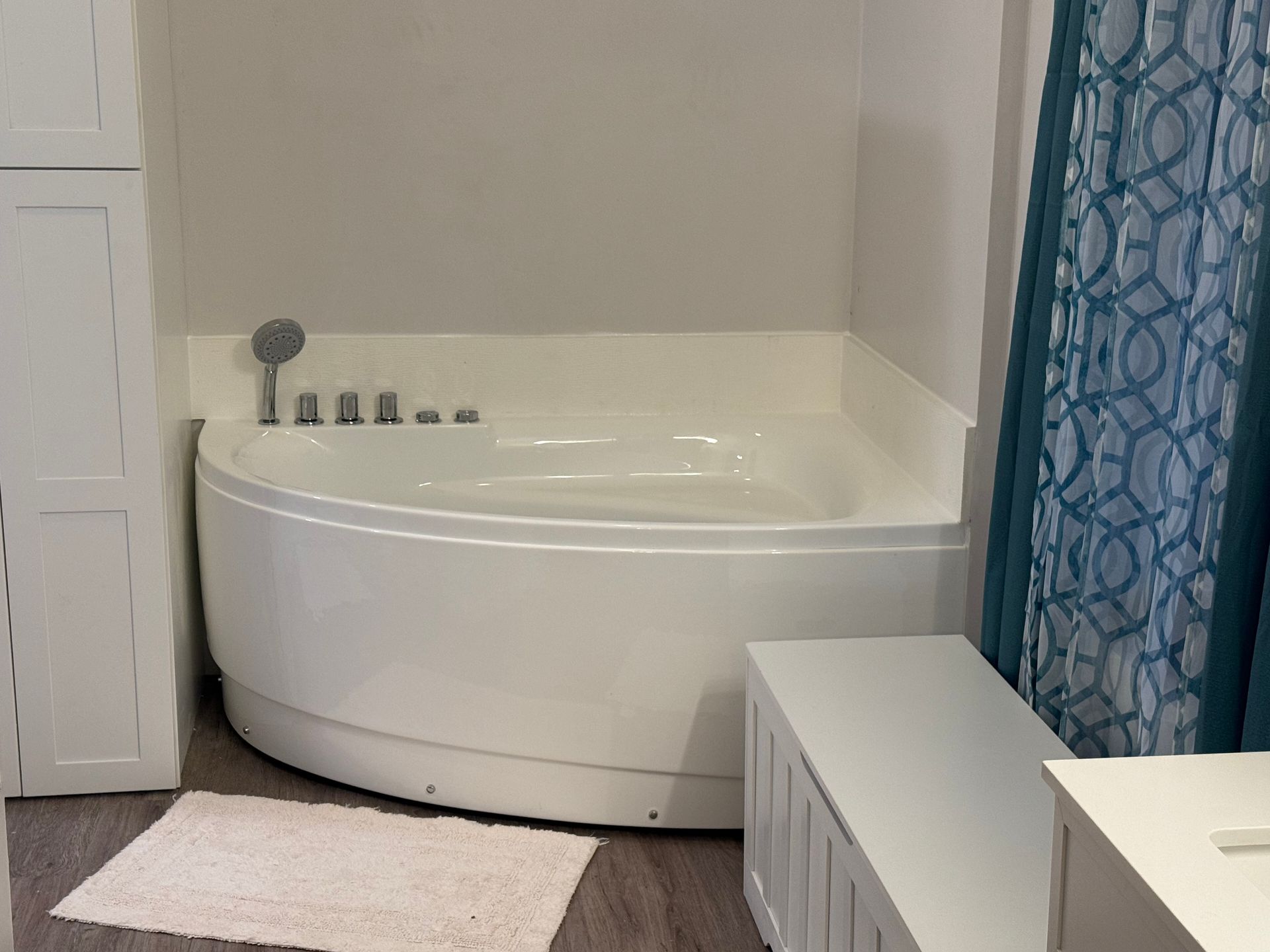 Small white Fibo corner bathtub in a bathroom with a blue shower curtain and white bench.