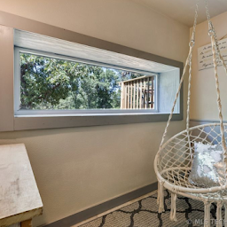 A white macramé swing chair sits in a room with a wide, rectangular window looking out onto a wooden deck and trees.