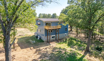 A blue, two-story house with a wooden deck stands in a sunny, tree-filled, wooded landscape.