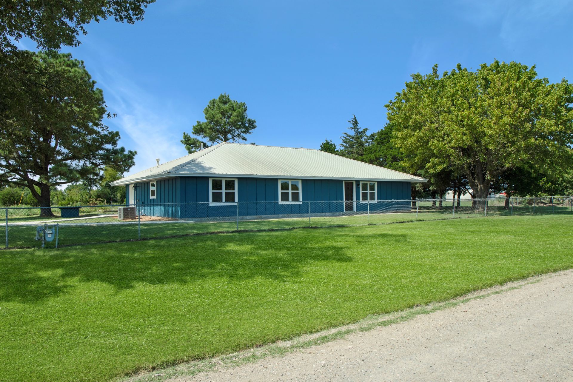 A blue, one-story house with a grey roof sits on a large grassy lot under a clear blue sky.