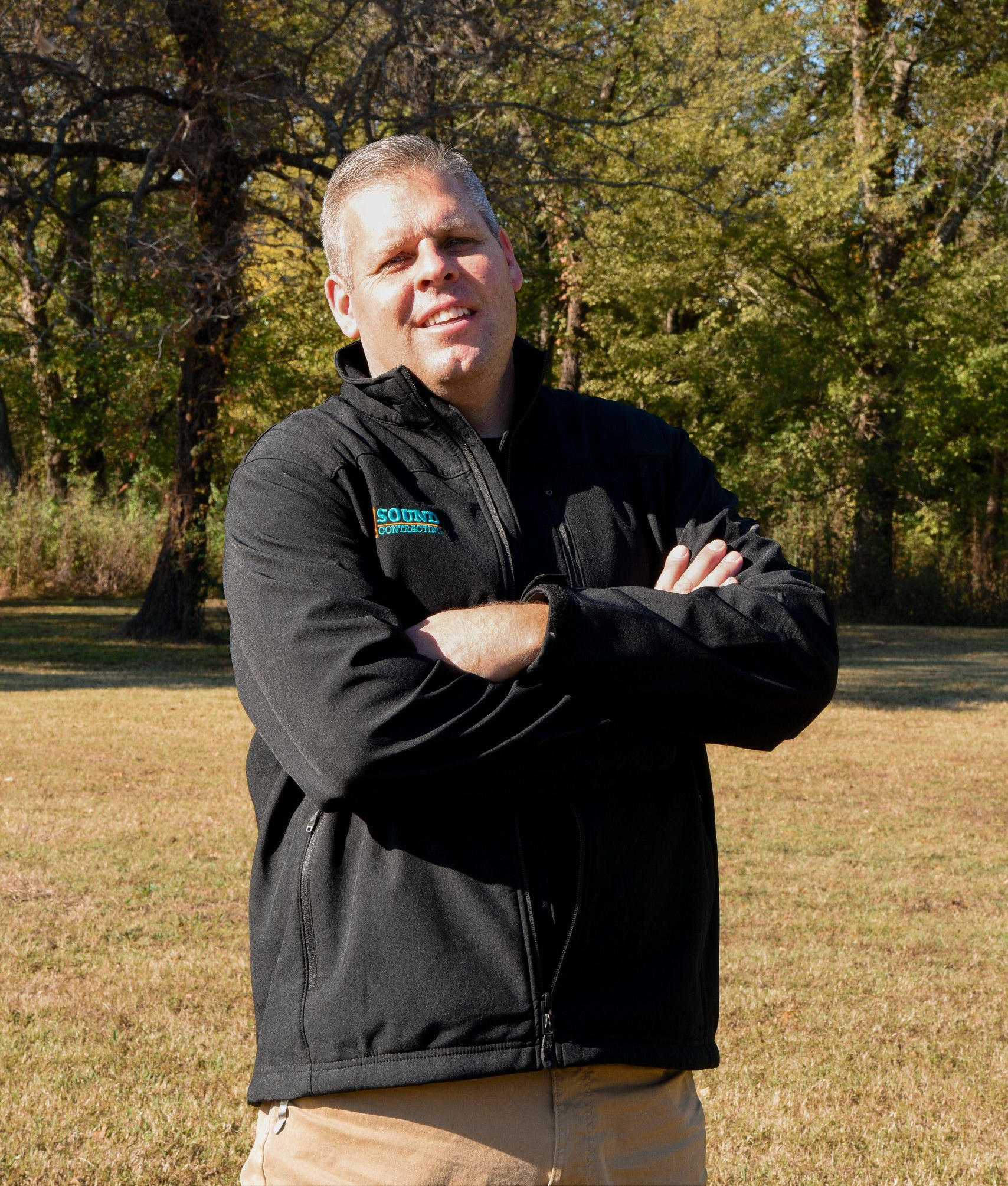 A person with a smiling expression stands with arms crossed in a field, wearing a black jacket and tan pants.