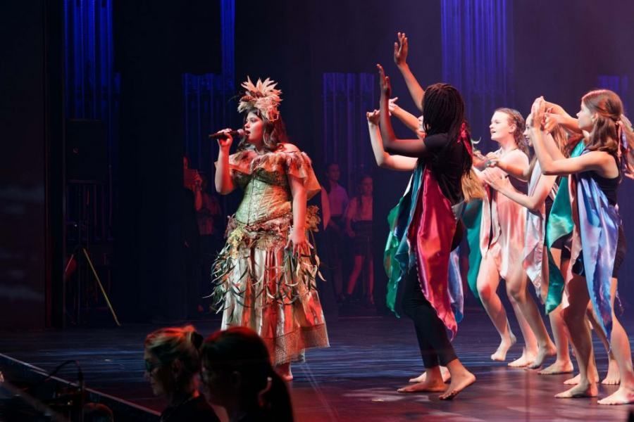 A woman is singing into a microphone while a group of young girls are dancing on a stage.