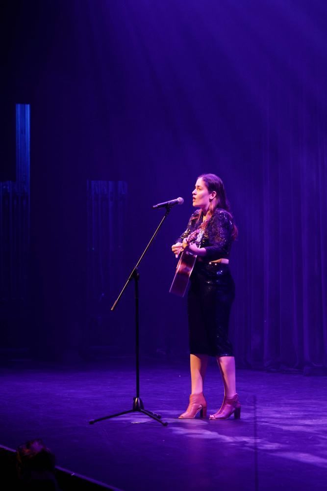 A woman is singing into a microphone on a stage while holding a guitar.