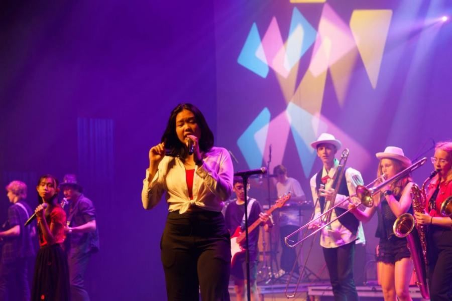 A woman is singing into a microphone on a stage with a group of people playing instruments.
