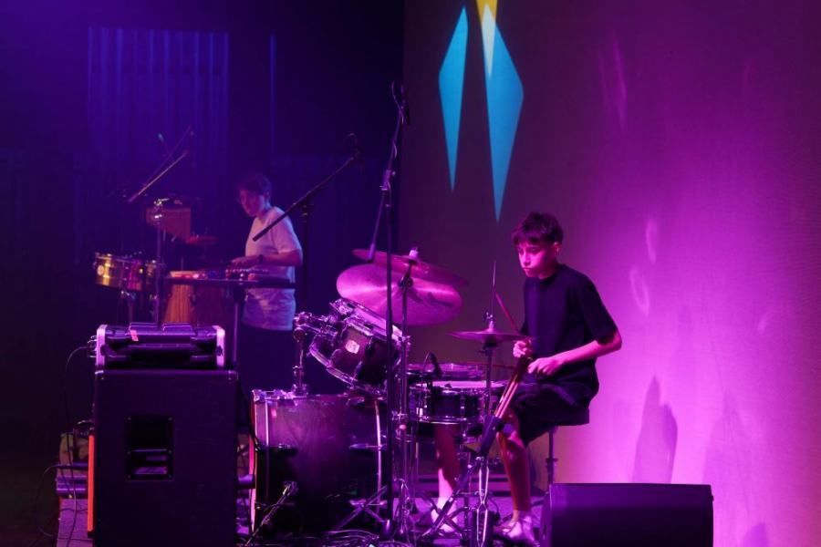 A man is playing drums on a stage with purple lights behind him.