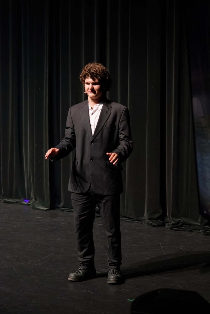 A man in a suit is standing on a stage in front of a black curtain.