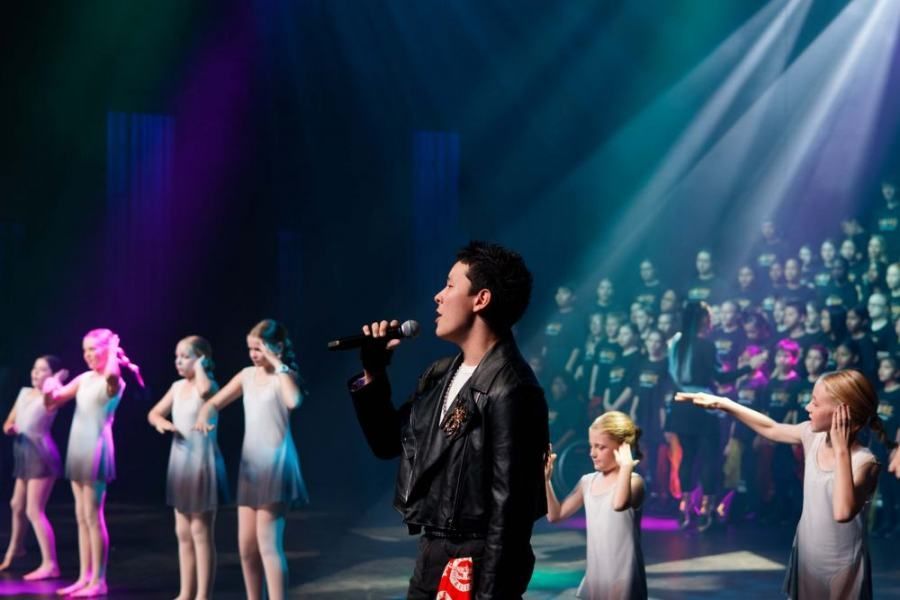 A man is singing into a microphone on a stage with a group of children.