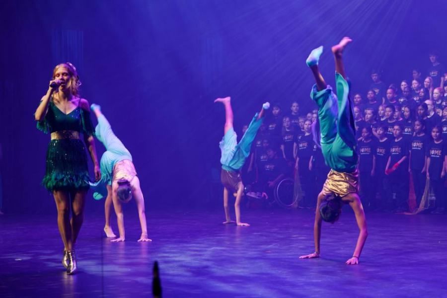 A group of children are doing handstands on a stage while a woman sings into a microphone.