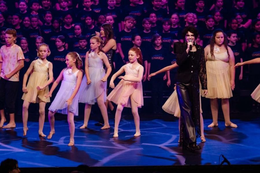 A group of young girls are dancing on a stage with a man singing into a microphone