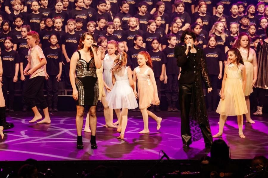 A group of children are standing on a stage with a woman singing into a microphone.