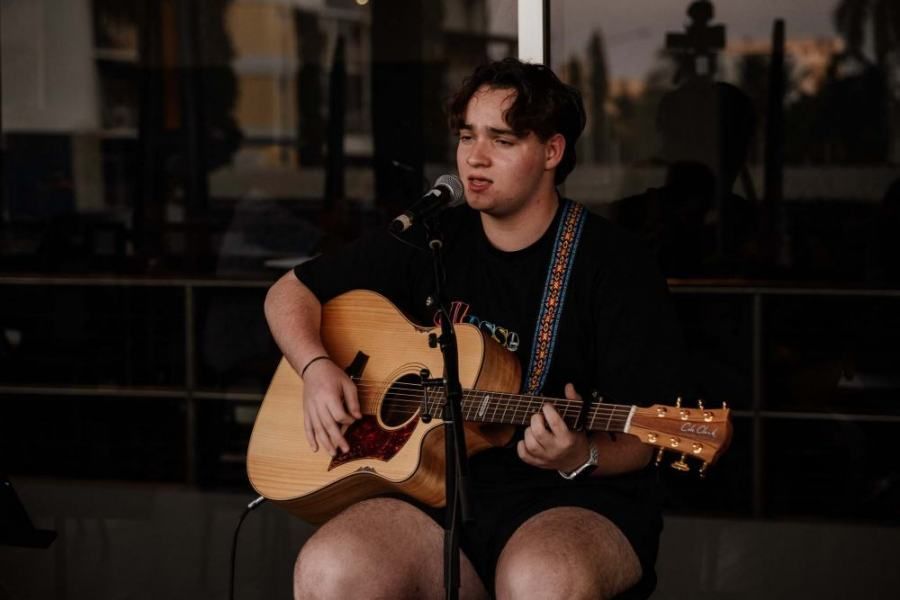 A young man is sitting on a chair playing a guitar and singing into a microphone.
