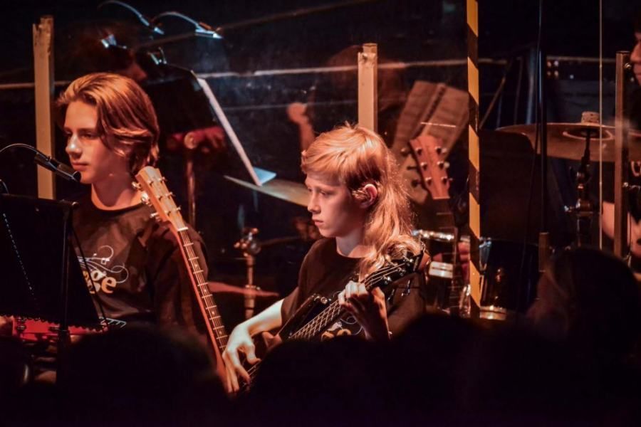 Two young men are playing guitars on a stage in front of an orchestra.