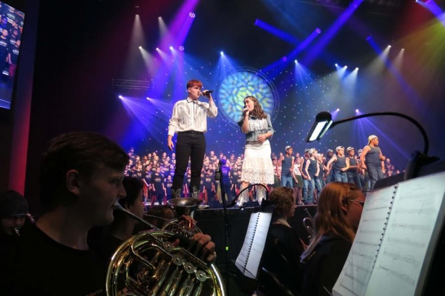 A man and a woman are singing on a stage in front of an orchestra.
