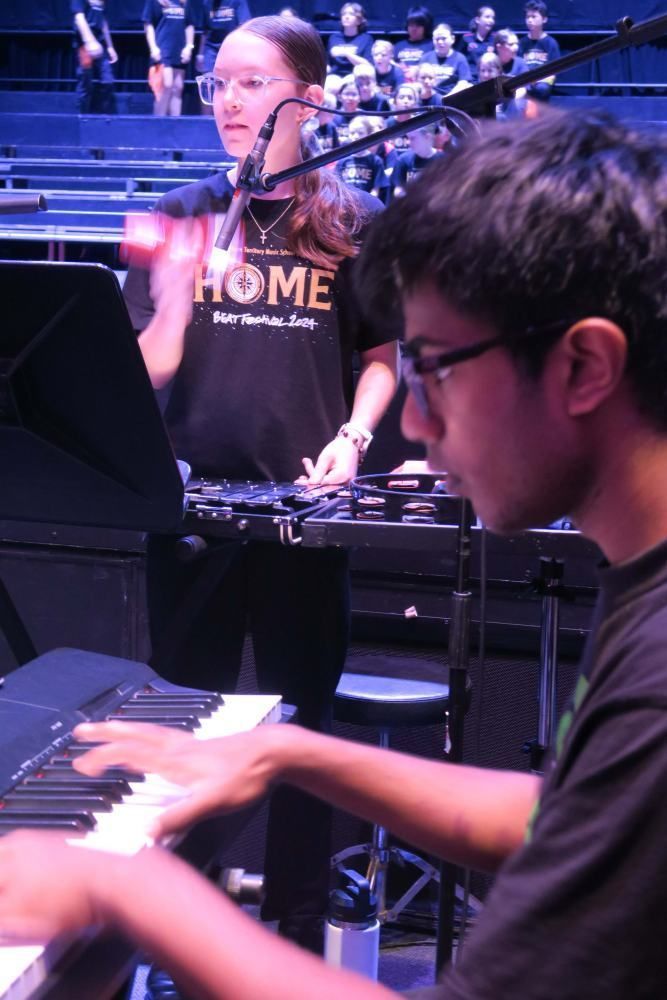 A man playing a keyboard with a woman behind him wearing a shirt that says home